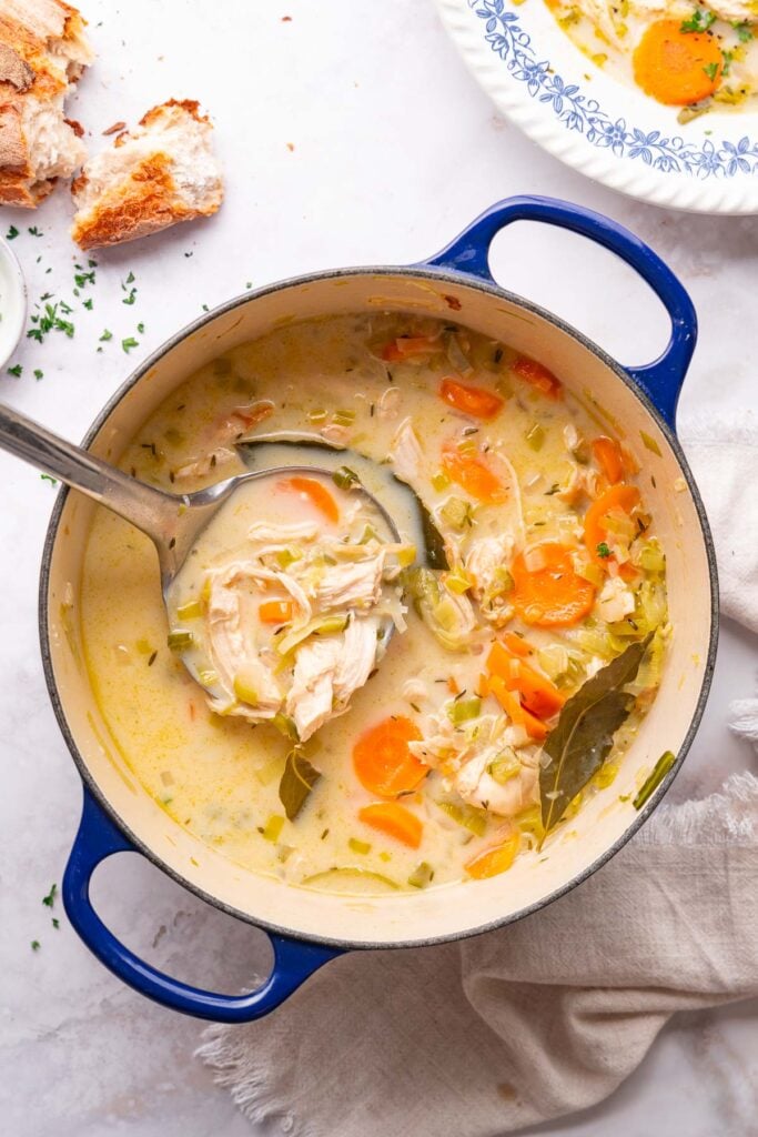 A pot of creamy vegetable chicken soup, filled with tender chicken, carrots, leeks and fragrant bay leaves. A ladle rests on the rim of the pot. Nearby, a plate holds even more of this creamy delight and a piece of bread ready to accompany it.  