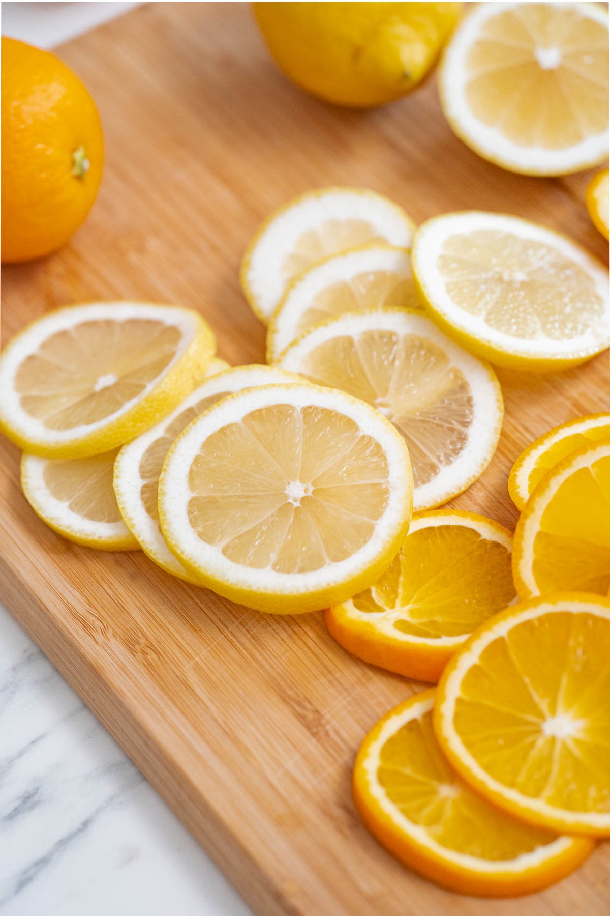 Sliced lemons and oranges on a wooden cutting board with whole fruit in the background.