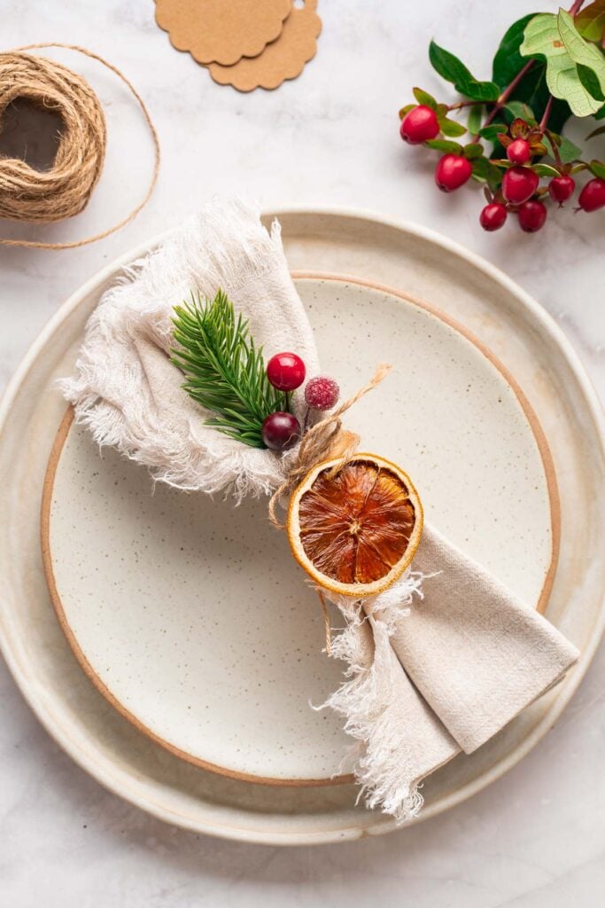 Festive table decoration with a beige napkin tied with twine, adorned with a slice of dried orange, greenery and red berries on a white plate. The string and berries are in the background. 