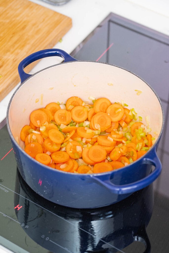 Une casserole bleue sur la cuisinière contient des carottes tranchées et des oignons hachés sautés, formant la base aromatique d'une copieuse soupe de haricots blancs et de tomates.