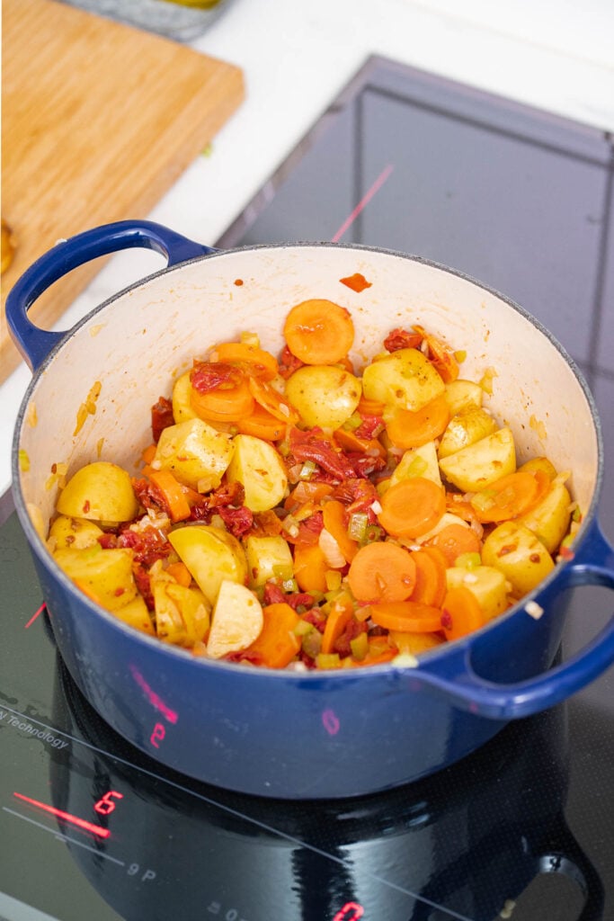 Une casserole bleue sur une cuisinière contient des pommes de terre hachées, des carottes, des tomates et des épices.