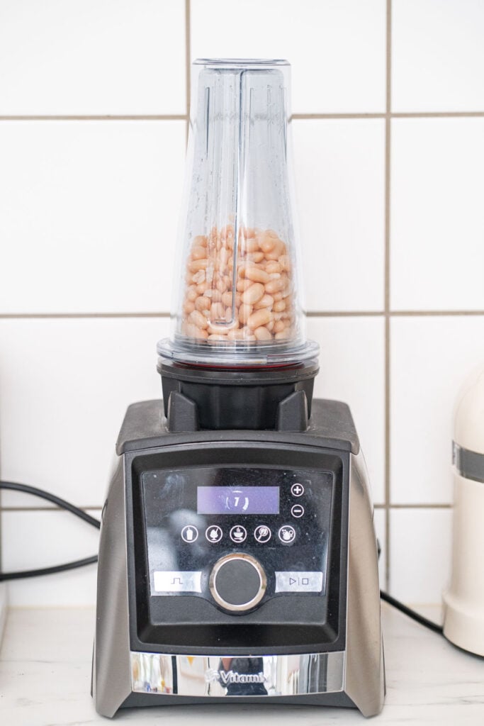 Un mixeur posé sur un comptoir de cuisine contient des haricots dans son récipient transparent, posé sur un fond de mur carrelé.