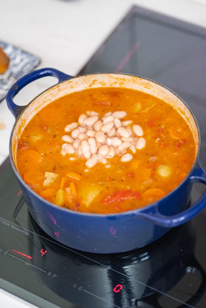 Une marmite bleue remplie de soupe aux haricots blancs et aux tomates est posée sur une table de cuisson à induction, présentant un mélange copieux de soupe aux légumes et de haricots blancs.