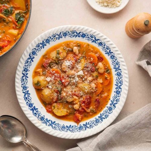 A bowl of vegetable stew with beans and zucchini, reminiscent of a hearty white bean and tomato soup, is topped with grated cheese on a decorative plate. Nearby are a napkin, spoon, fork and a small plate of grated cheese.