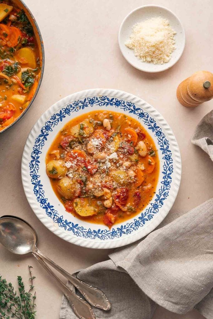 Un bol de ragoût de légumes avec des haricots et des courgettes, rappelant une soupe copieuse aux haricots blancs et aux tomates, est garni de fromage râpé sur une assiette décorative. À proximité se trouvent une serviette, une cuillère, une fourchette et une petite assiette de fromage râpé.