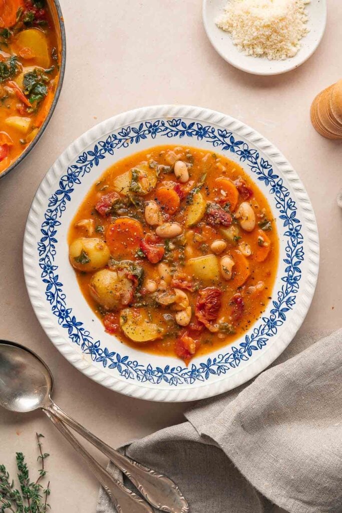 Un bol de soupe haricots blancs tomates est servi dans un plat décoratif bleu et blanc, accompagné d'une cuillère et d'une petite assiette de fromage râpé.