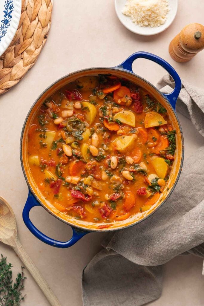 Une marmite bleue remplie d'une soupe copieuse de haricots blancs et de tomates, débordant de haricots, de pommes de terre, de carottes et de légumes verts. Elle repose sur une surface beige à côté d'une cuillère en bois et d'un petit bol de fromage râpé.