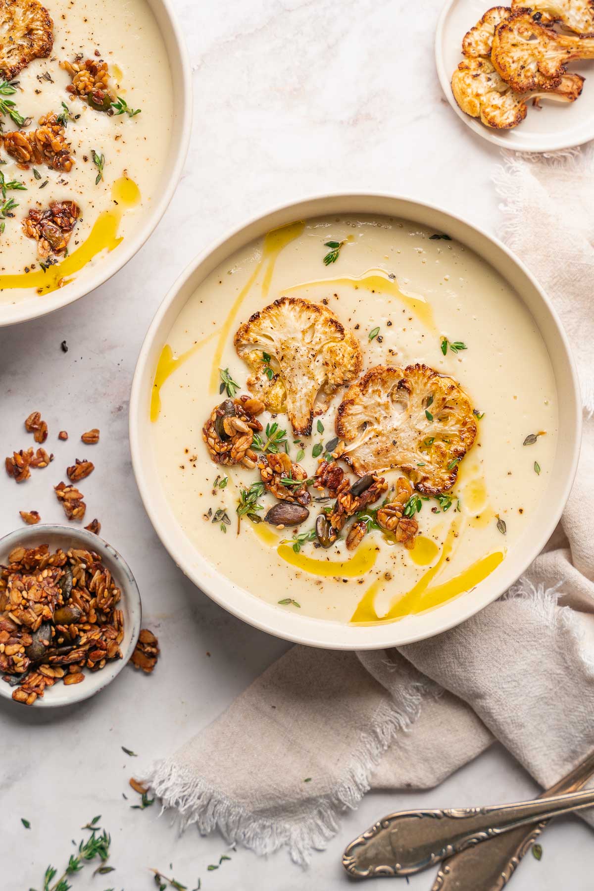 Un bol de velouté de chou-fleur sans pomme de terre, garni de chou-fleur rôti, d'herbes et de graines, repose sur une surface claire avec une serviette et une cuillère à proximité.