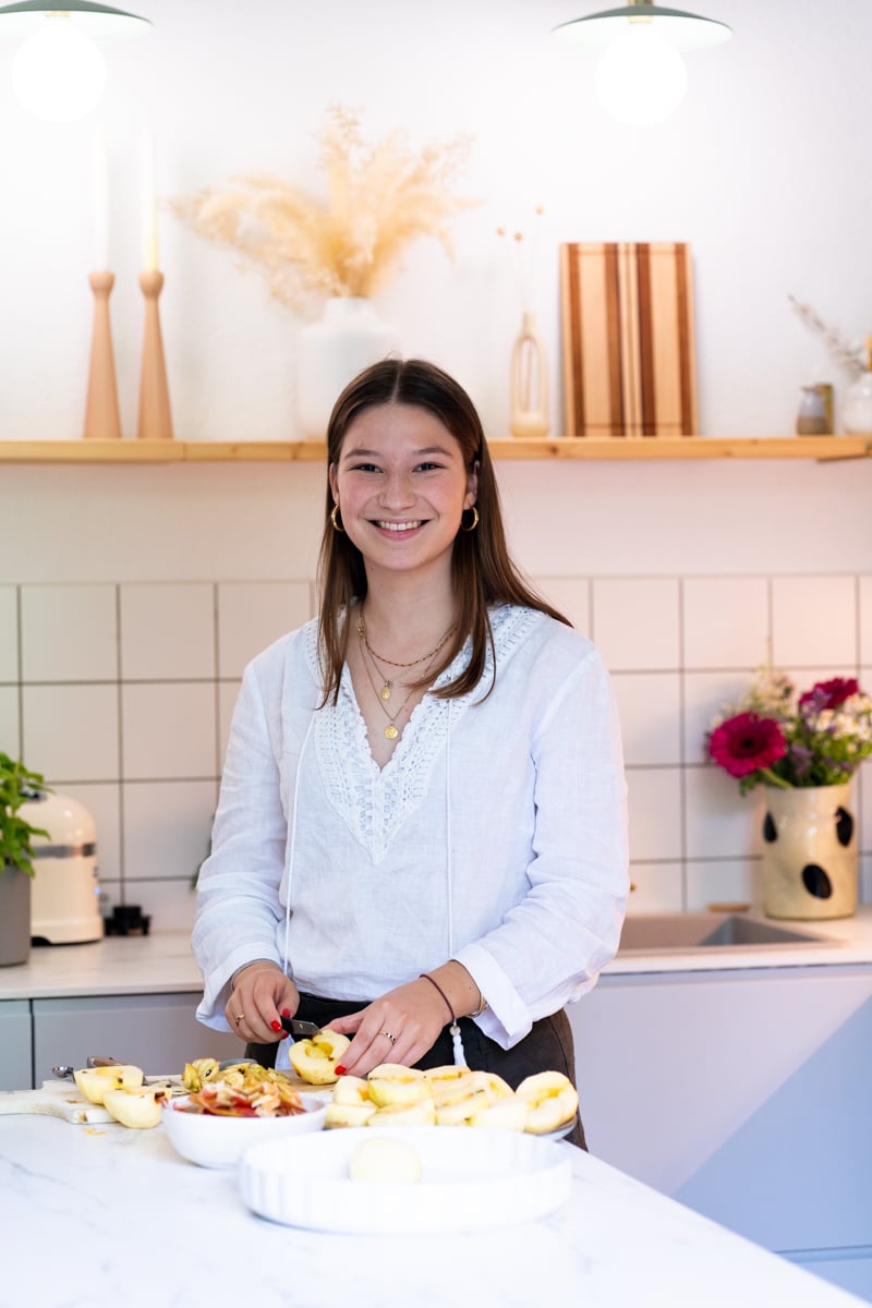 A woman in a kitchen, featured on a popular food blog, is cutting apples on a counter. She's wearing a white blouse and smiling warmly. The kitchen features tiled walls, a decorated shelf and a vase with flowers, creating a welcoming atmosphere.  