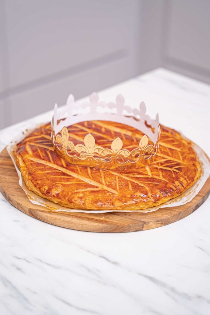 A golden-brown Galette des Rois, topped with a decorative paper crown, rests on a round wooden board on a marble surface, a delicious nod to French Tradition.