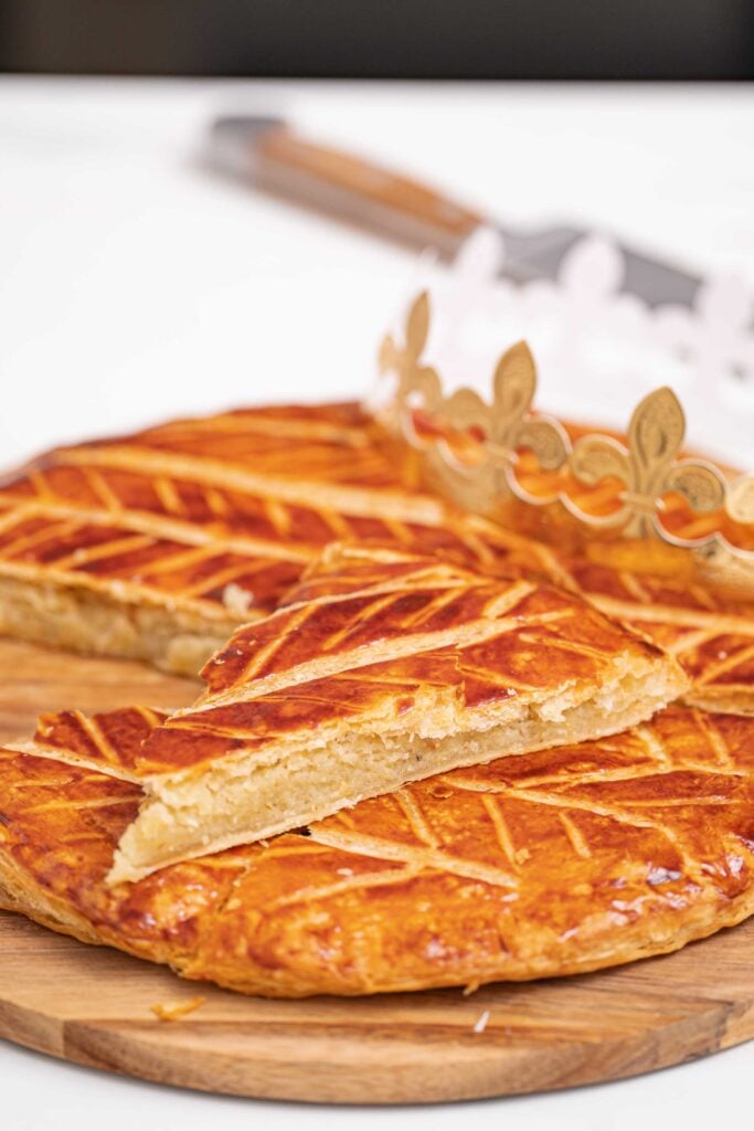 Golden Galette des Rois with an intricate pattern on top, a slice cut out, set on a wooden board. I love the way a decorative paper wreath sits proudly in the background, capturing the essence of French Tradition. 