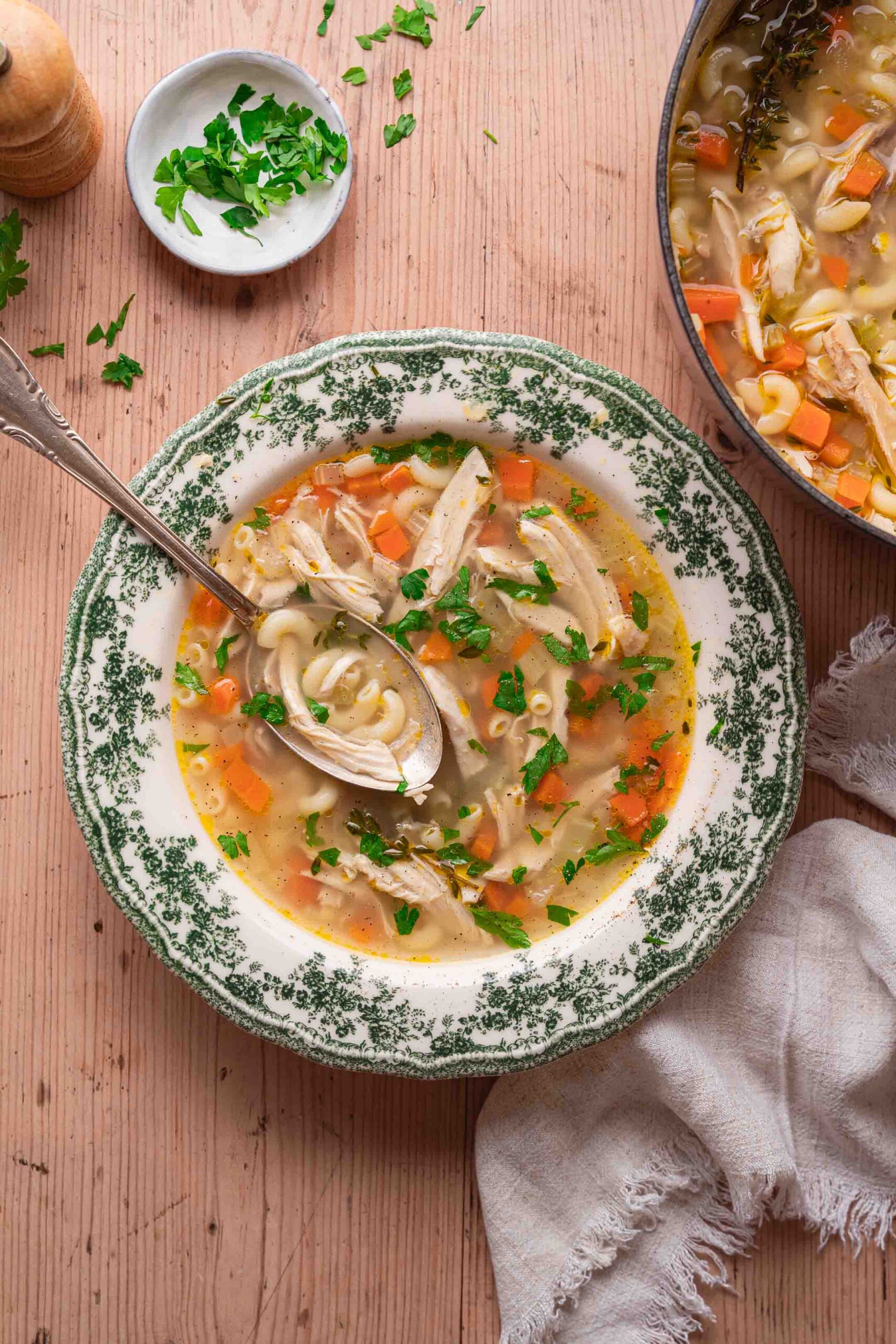 A bowl of chicken-vegetable pasta soup with carrots, parsley and a spoon rests on a wooden table. A cloth napkin and an extra portion of parsley lie nearby, ready to enhance this comforting dish. 