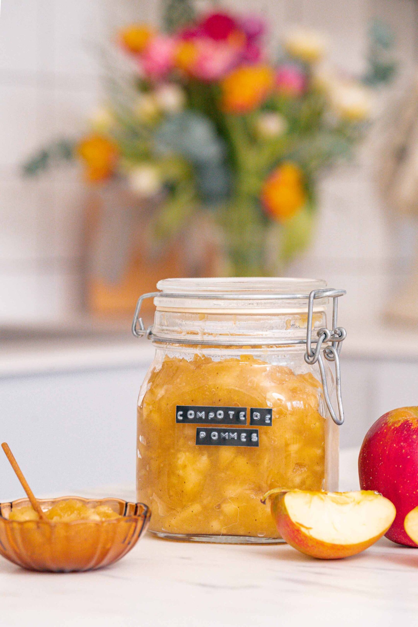 Un bocal étiqueté « Compote de Pommes Maison », rempli de compote de pommes maison, est posé sur un comptoir de cuisine, à côté d'une petite assiette et de tranches de pomme. Une composition florale floue ajoute du charme à l'arrière-plan.
