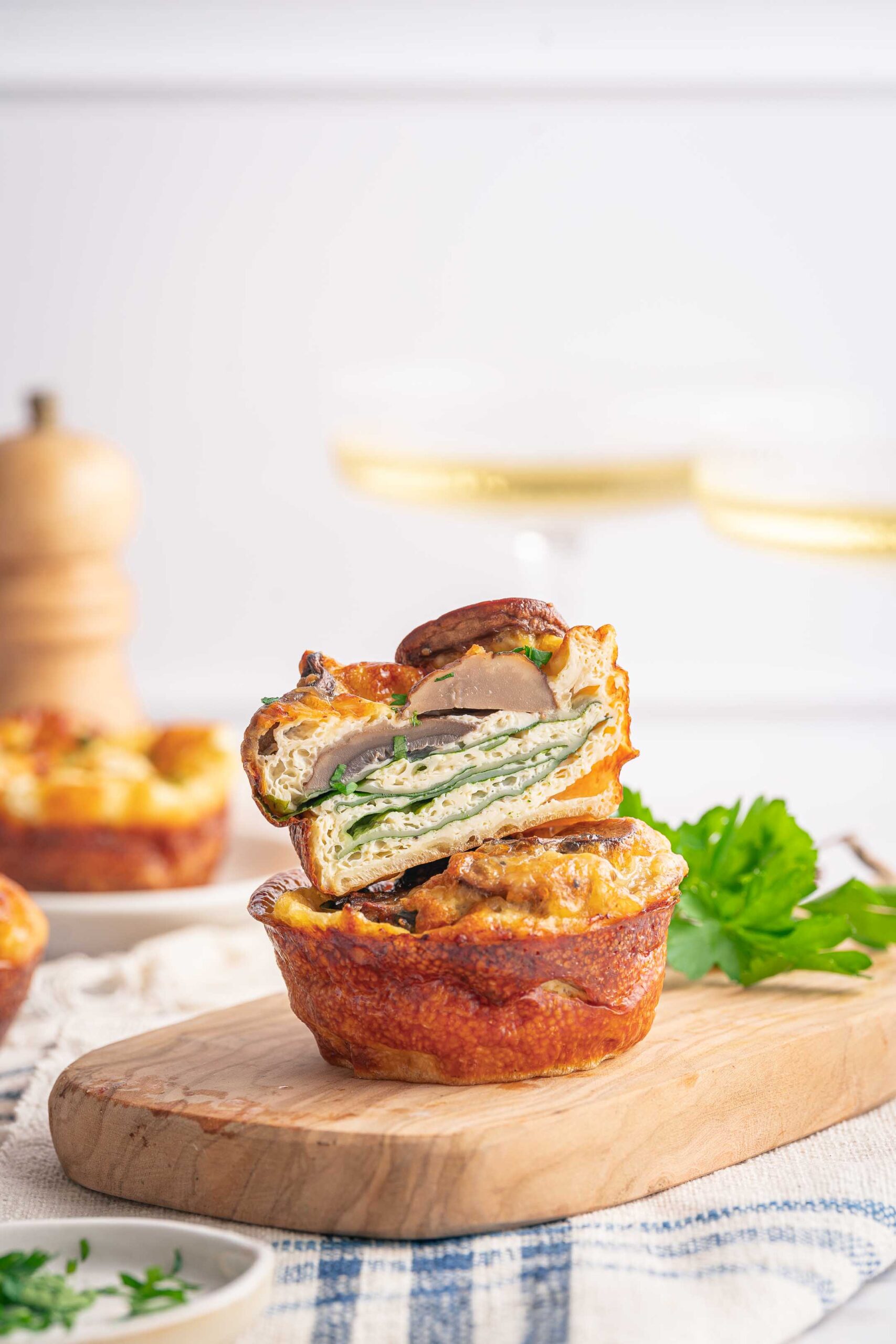 Un muffin salé tranché, rappelant une mini-quiche sans croûte, garni d'épinards et de champignons, repose sur une planche de bois, agrémentée de persil. En arrière-plan flou, d'autres muffins et un moulin à poivre pour un assaisonnement parfait.