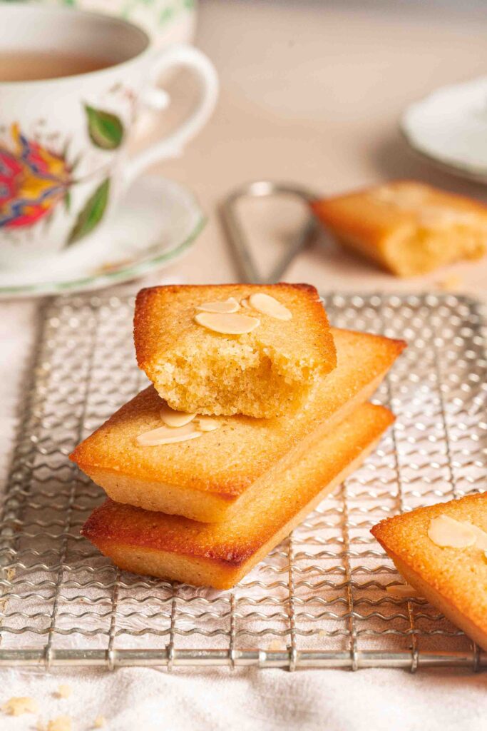 Trois financiers rectangulaires aux amandes sont empilés sur une grille de refroidissement, dont un partiellement dévoré. Une tasse de thé et d'autres financiers sont visibles en arrière-plan.