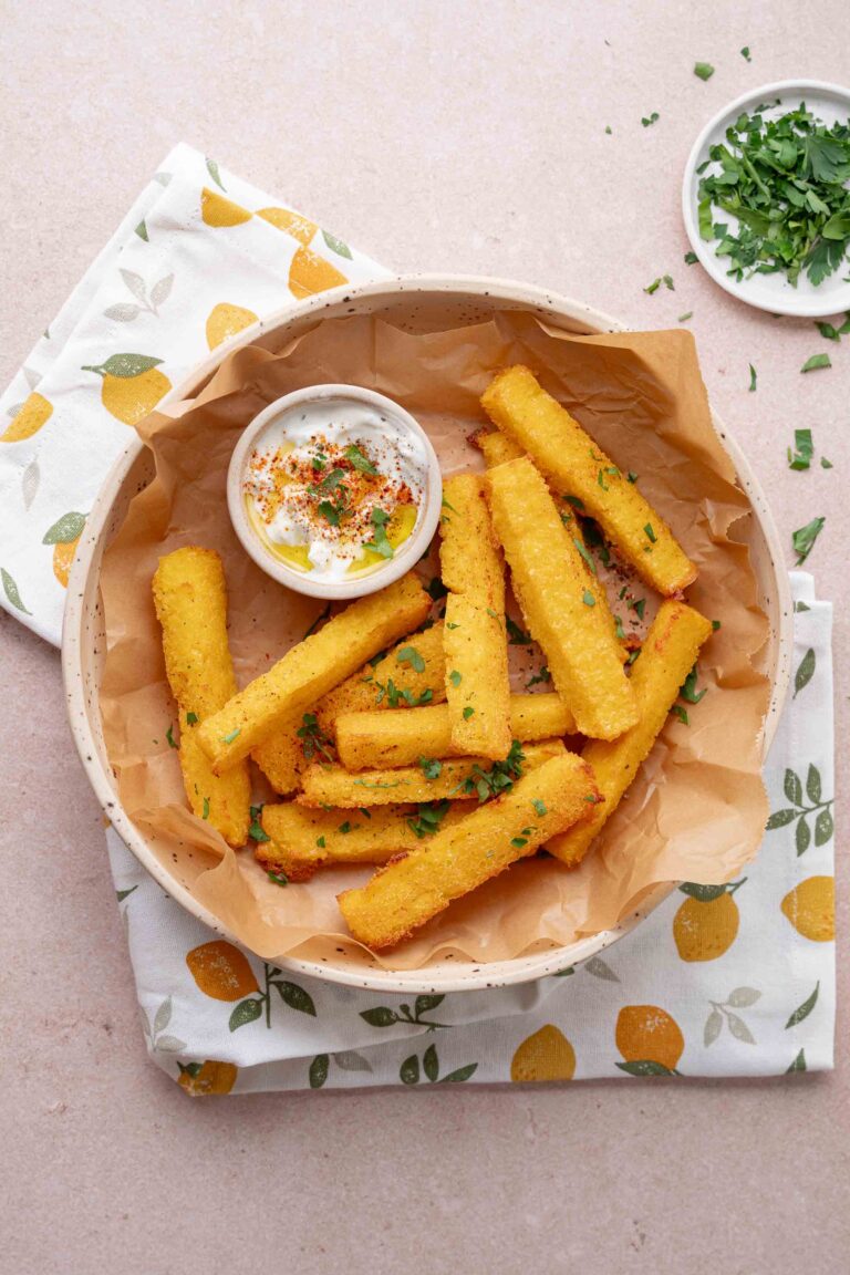 Un bol de frites de polenta dorées avec un petit plat de trempette, garni d'herbes hachées, sur une serviette à motifs citron.