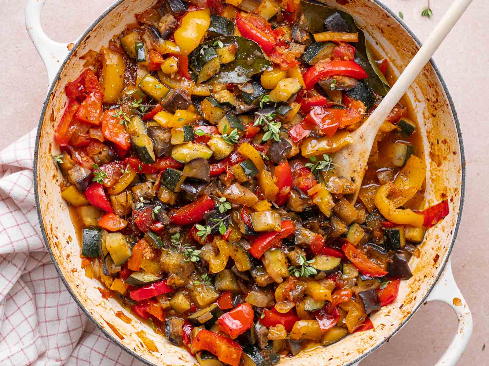 A white pan filled with cooked ratatouille, featuring chopped zucchini, eggplant, red and yellow bell peppers, and garnished with fresh herbs illustrates a classic Ratatouille recipe. A wooden spoon rests in the pan.