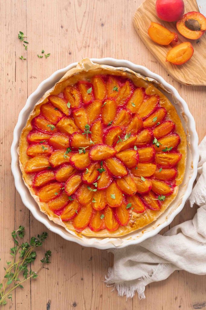 A tart baked in a white dish is garnished with carefully arranged slices of orange apricot. Fresh herbs and a halved apricot are placed on a wooden surface.