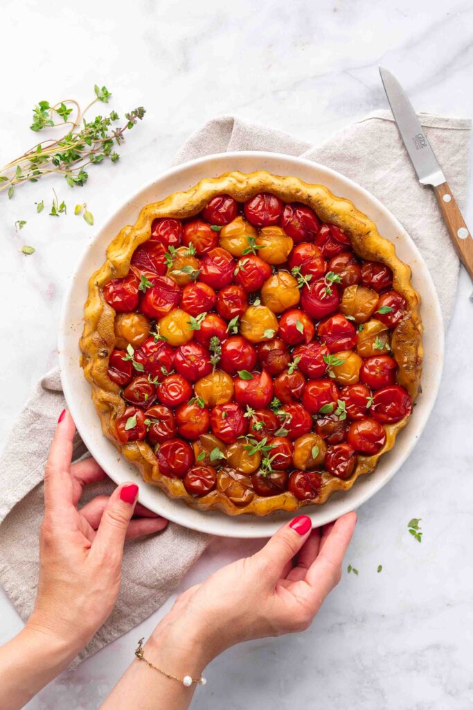 One person holds a plate containing a baked tomato tart, garnished with red and yellow cherry tomatoes and fresh herbs, placed on a marble surface, with a knife and napkin nearby.