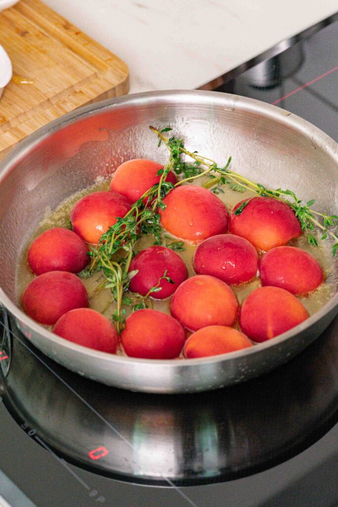 A stainless steel pan on the stovetop contains whole peaches and sprigs of fresh herbs simmering in the liquid.