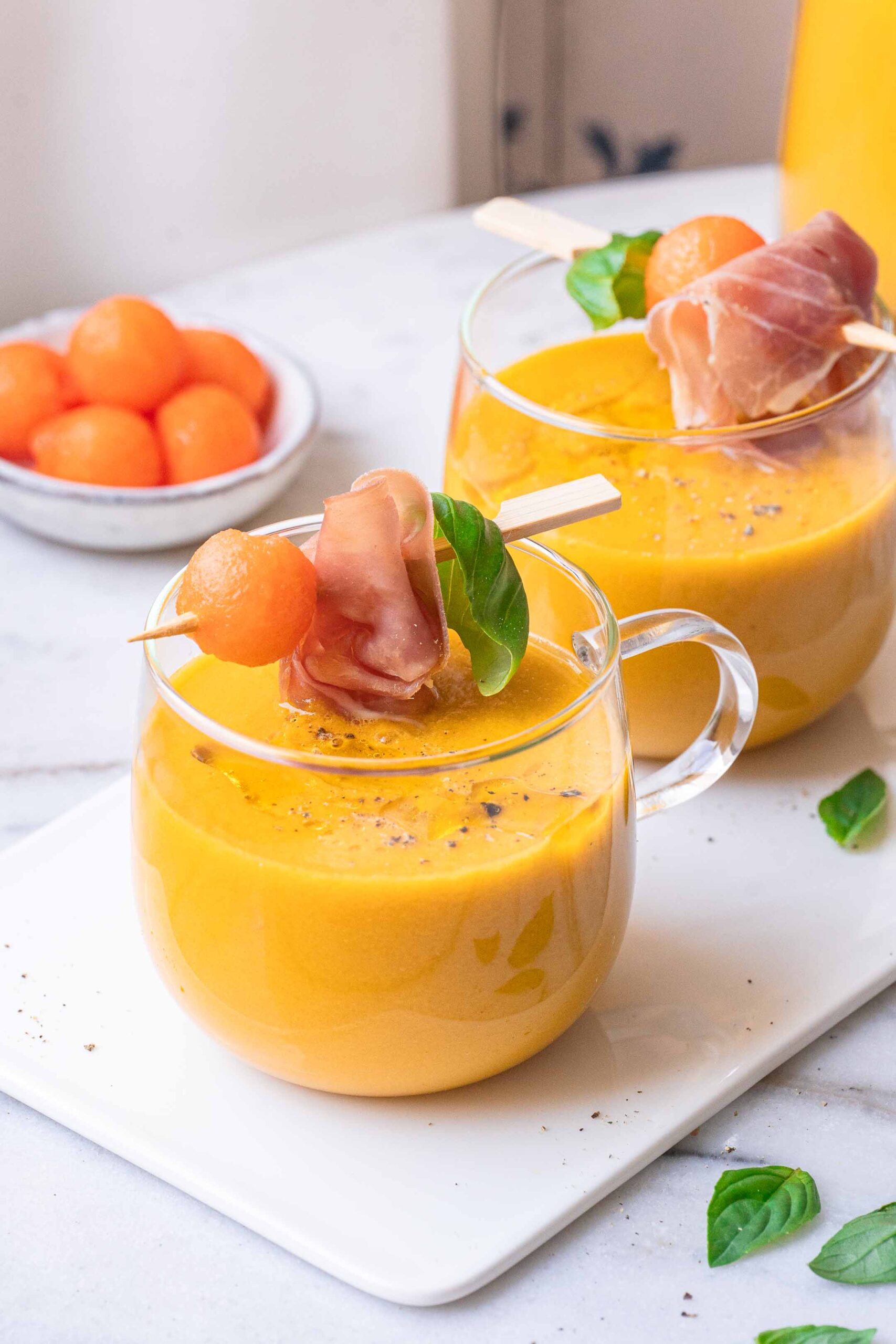 Two glasses of orange melon soup are garnished with prosciutto skewers, basil and melon balls. A bowl of melon balls is in the background. 