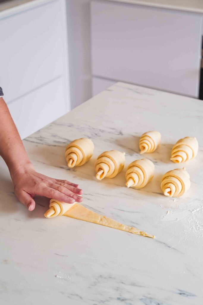 A person rolls out a piece of dough on a floured surface, with several unbaked croissants already formed in the background.