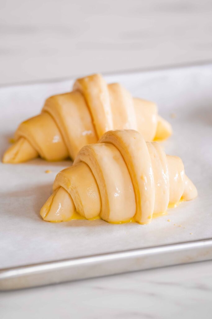 Two unbaked, egg-washed croissants are placed on a parchment-lined baking sheet.
