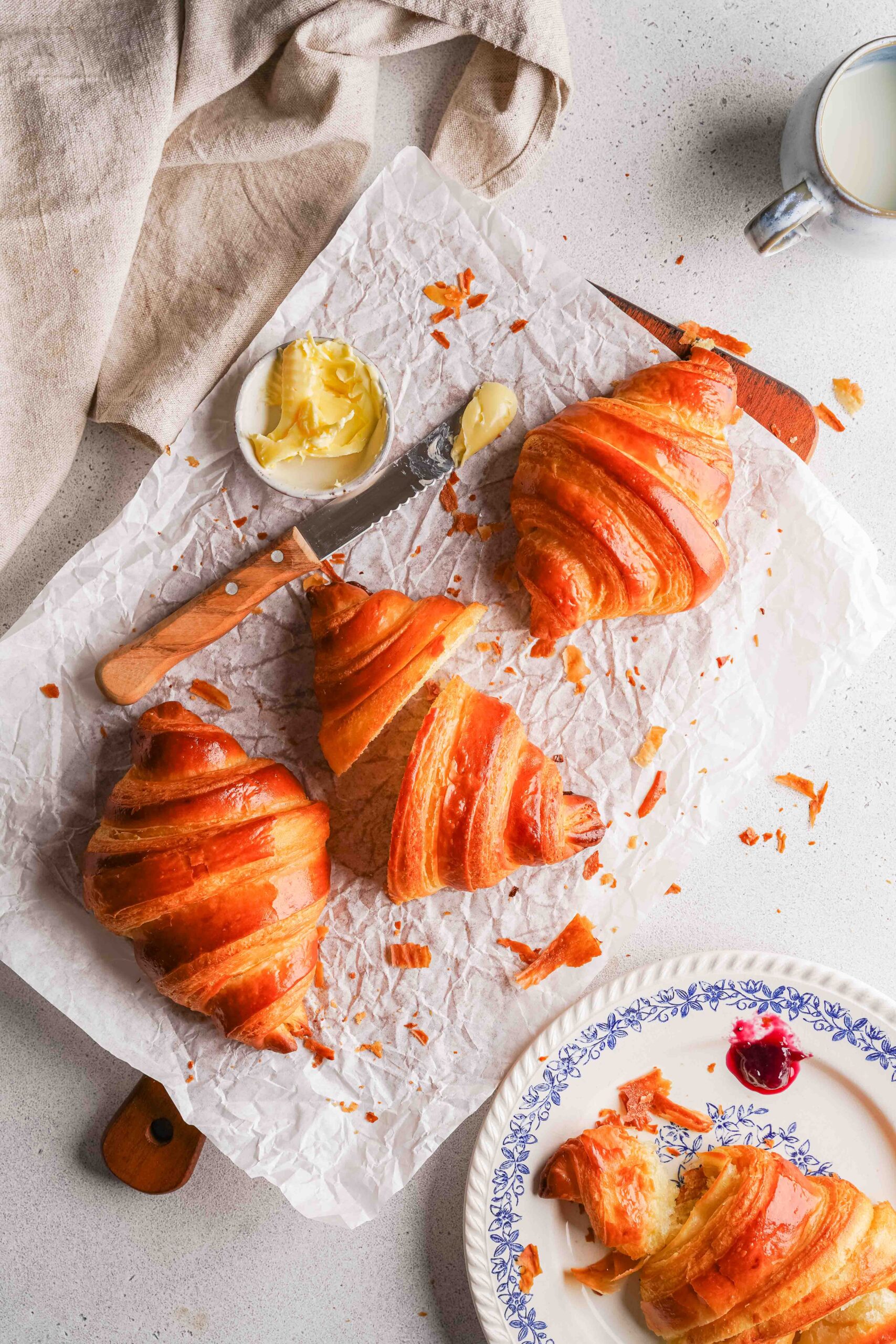 Trois croissants sur du papier sulfurisé avec un couteau et du beurre, à côté d'une assiette contenant un croissant partiellement mangé et une cuillerée de confiture rouge. Une tasse de lait se trouve à l'arrière-plan.