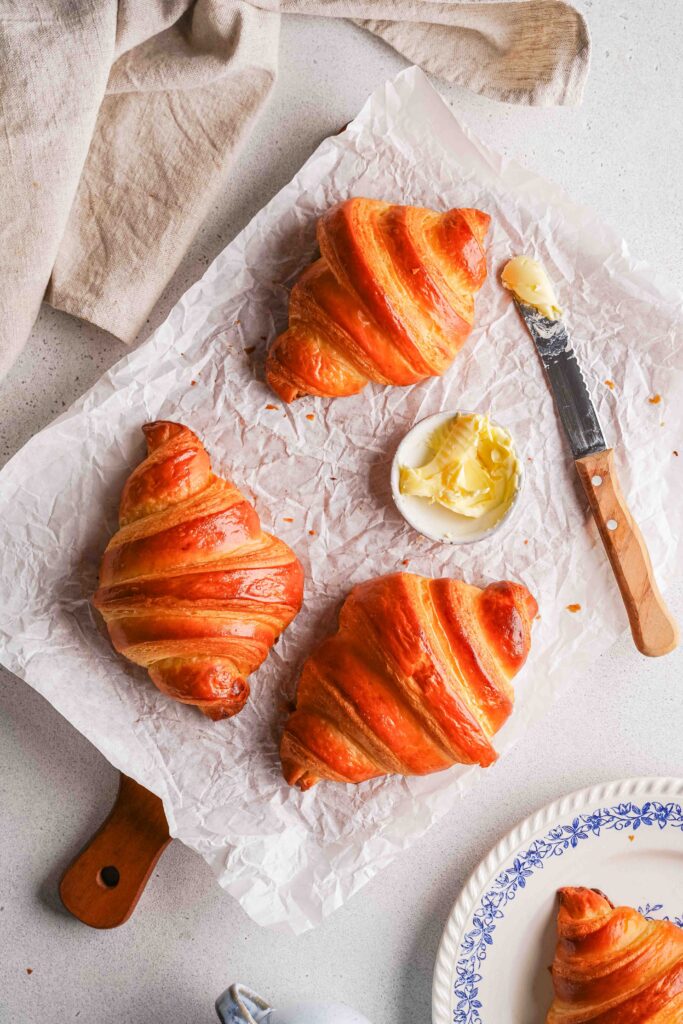 Three golden croissants sit on greaseproof paper, with a small dish of butter and a butter knife beside them; a beige cloth and a patterned plate are partially visible in the background.