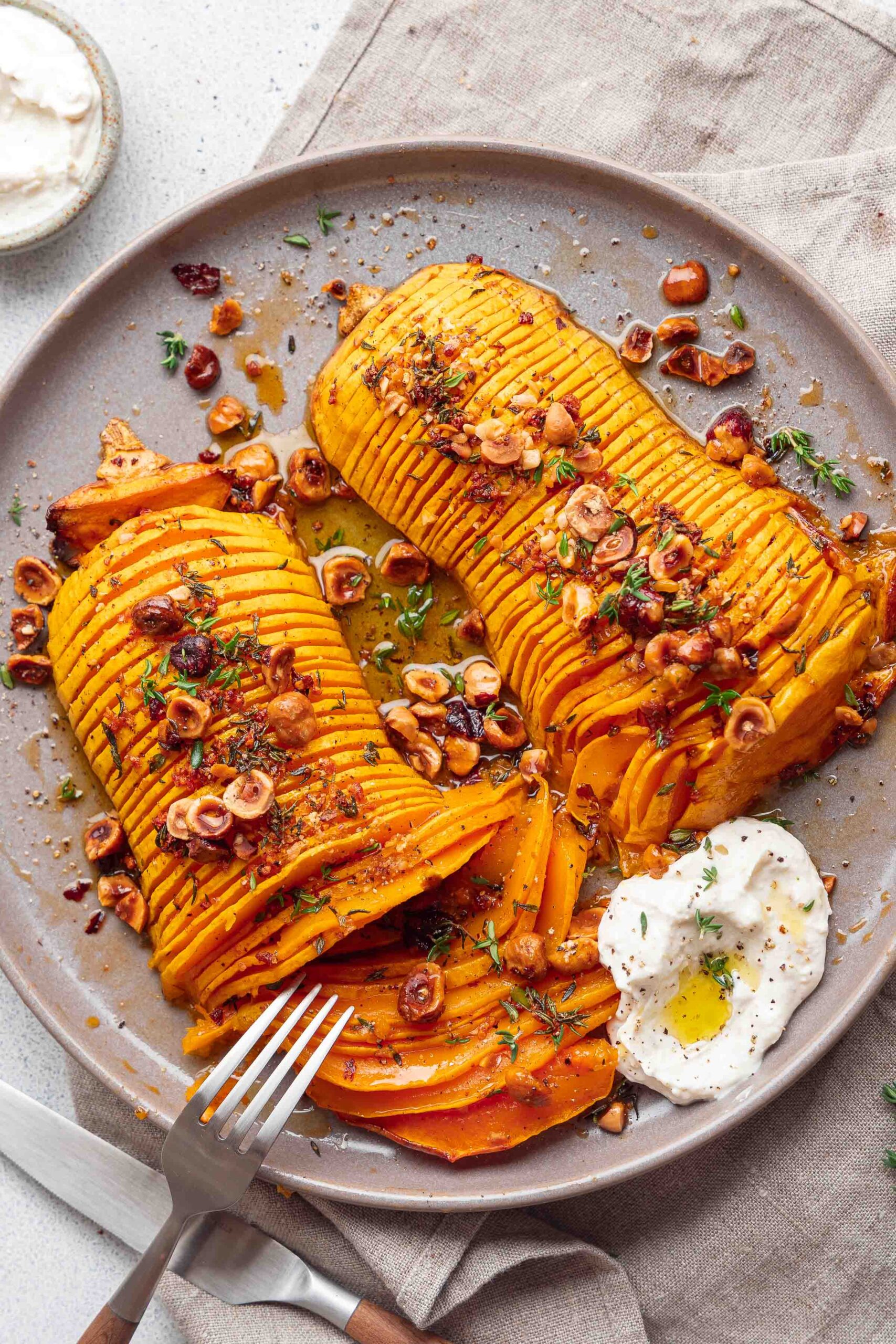 Gebratene Butternut-Kürbisscheiben mit gehackten Nüssen und Kräutern garniert, mit einem Löffel Joghurt und einem Spritzer Öl auf einem beigen Teller mit einer Gabel serviert.