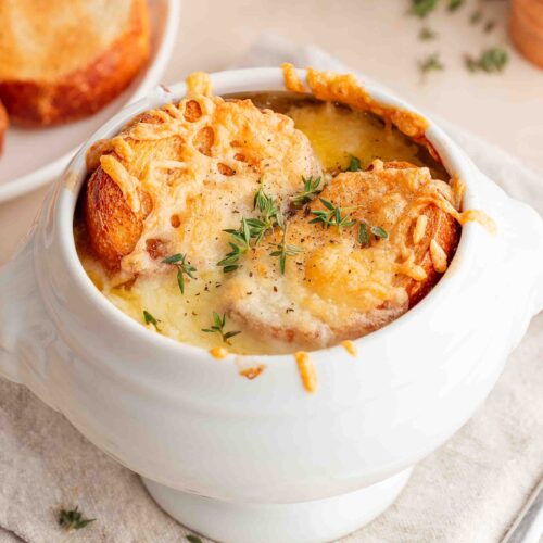 Eine weiße Keramikschüssel, gefüllt mit Zwiebelsuppe, garniert mit geröstetem Brot, geschmolzenem Käse und dekoriert mit frischem Thymian.