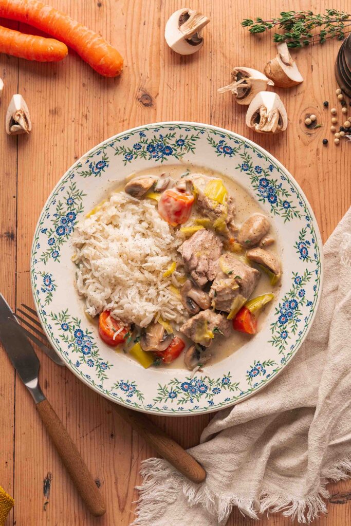 A plate of white rice and a creamy chicken stew with carrots, mushrooms and leeks, reminiscent of a French veal stew, garnished with herbs, on a wooden table with raw vegetables and cutlery next to it.