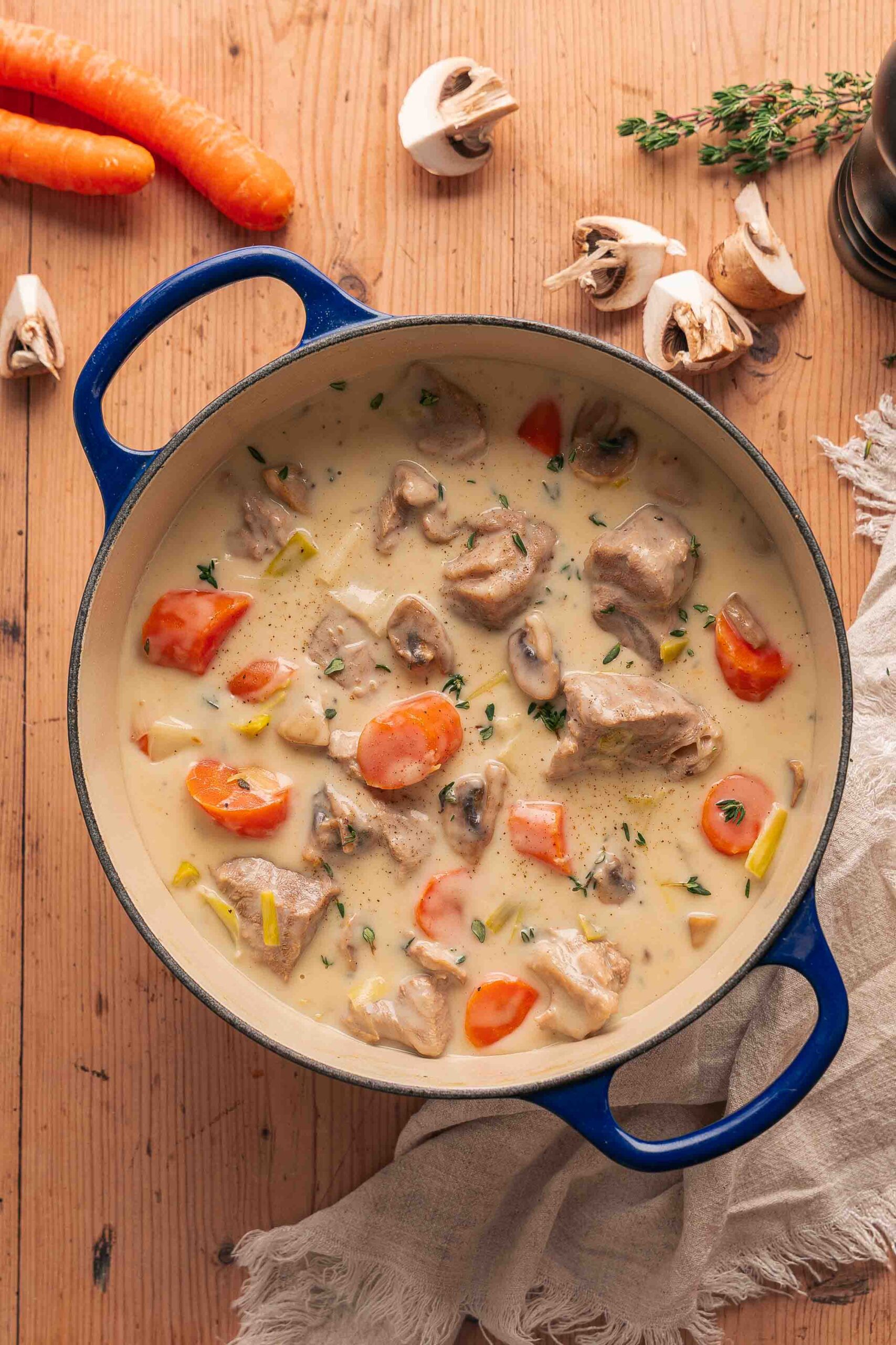 Une marmite bleue remplie de ragoût de veau français crémeux, avec de tendres morceaux de veau, des carottes émincées, des champignons et des herbes aromatiques sur une table en bois.
