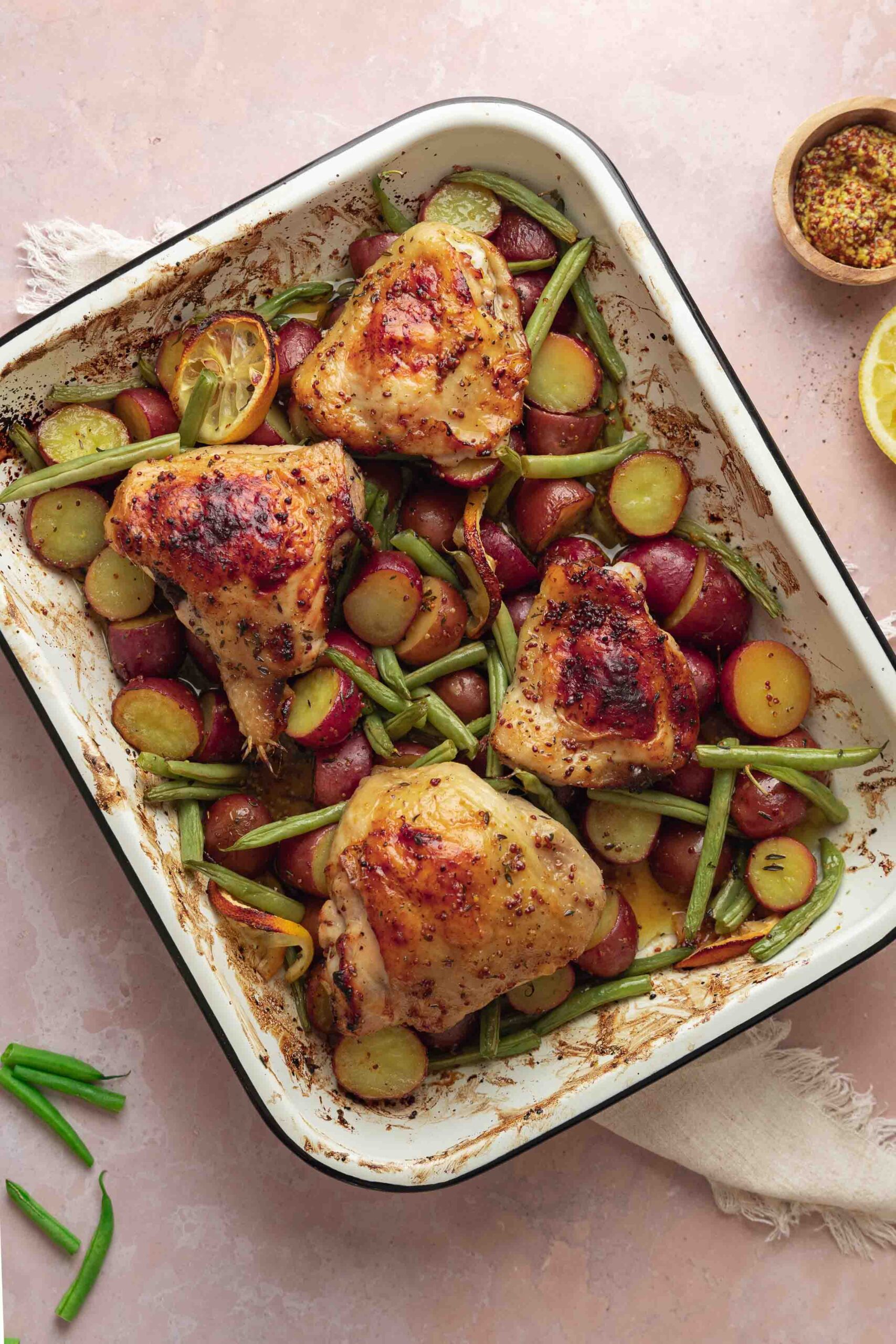 Gebackene Hähnchenschenkel mit gebratenen Kartoffeln und grünen Bohnen in einer weißen Auflaufform, garniert mit Zitronenscheiben, auf einer blassrosa Oberfläche.
