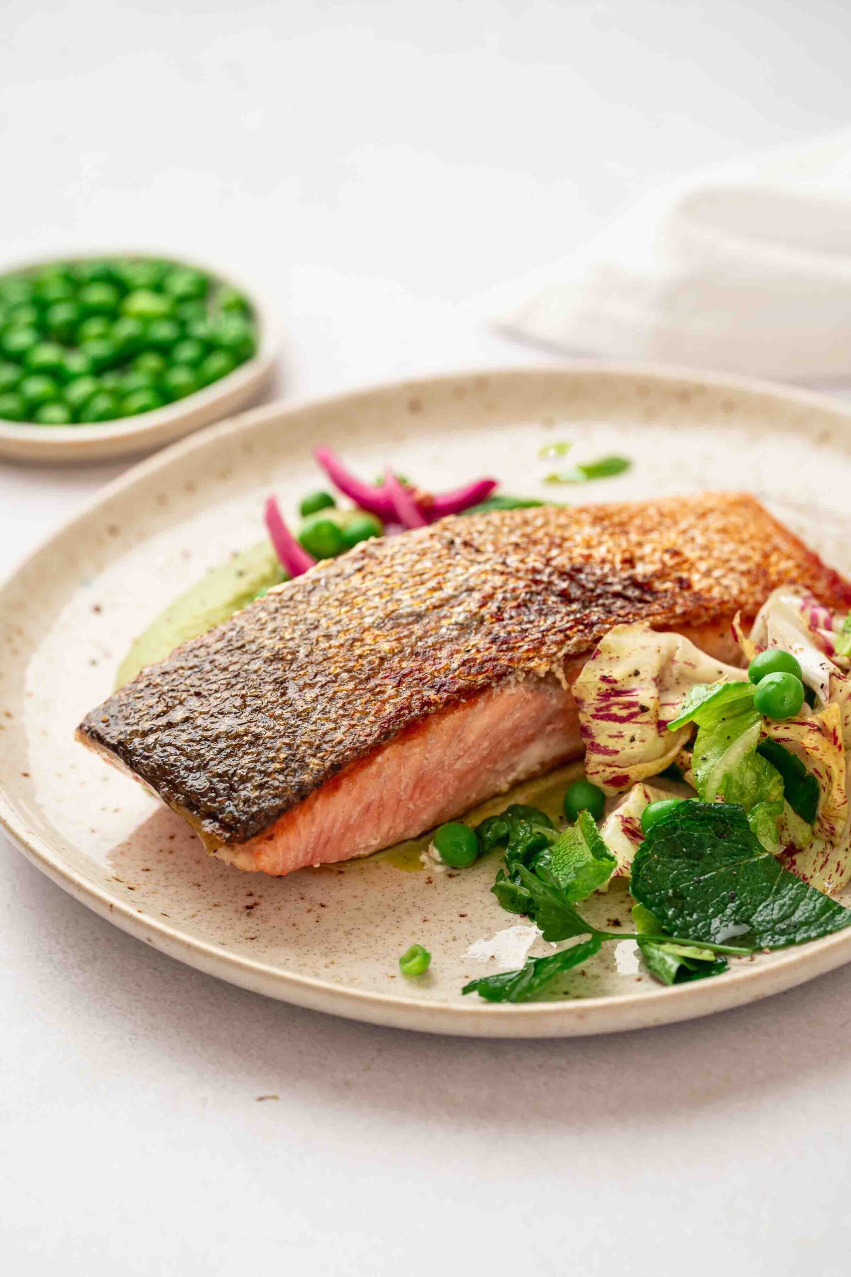 A plate with a crispy salmon steak, with crispy skin, served with green peas and sautéed vegetables. In the background is a bowl of pea puree. 
