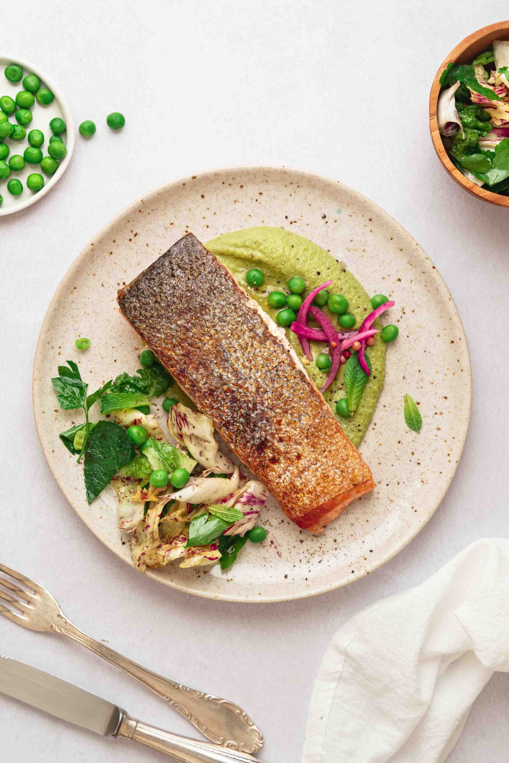 A plate with a crispy salmon steak, pea puree, mixed greens, peas and pickled onions. Next to it are a fork, a knife, a napkin and bowls of peas and salad. 