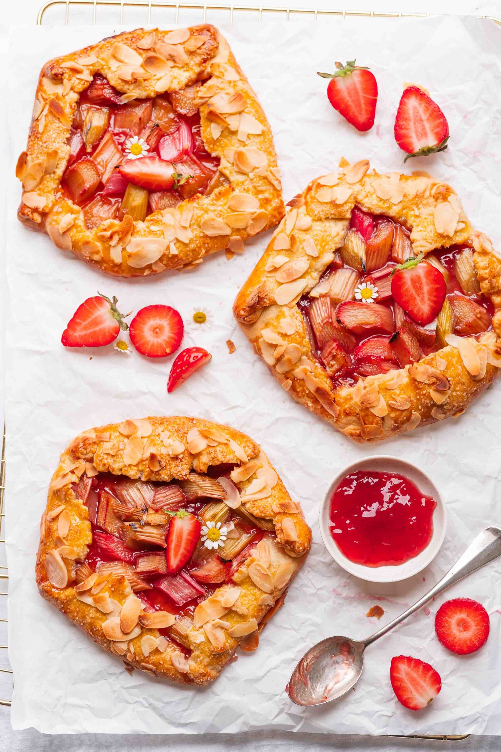 Drei Rhabarber-Erdbeer-Galettes mit Mandelscheiben werden auf Pergamentpapier angerichtet, umgeben von frischen Erdbeerhälften und einem Schälchen roter Marmelade mit einem Löffel.