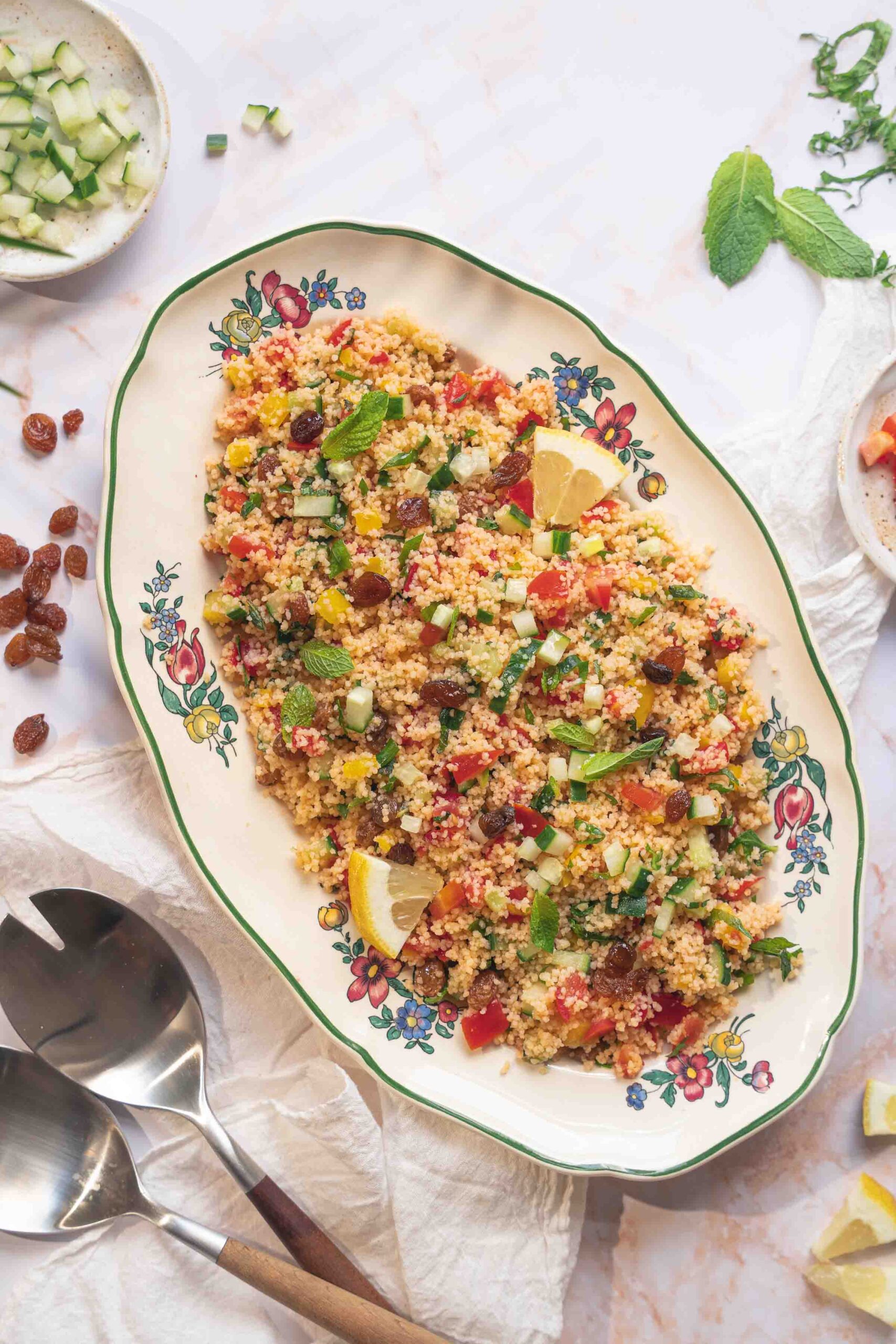 Un plat de salade de couscous aux légumes coupés en morceaux, aux raisins secs, aux feuilles de menthe et aux quartiers de citron — qui rappelle le taboulé —, servi dans un plat à motifs floraux, accompagné de couverts.