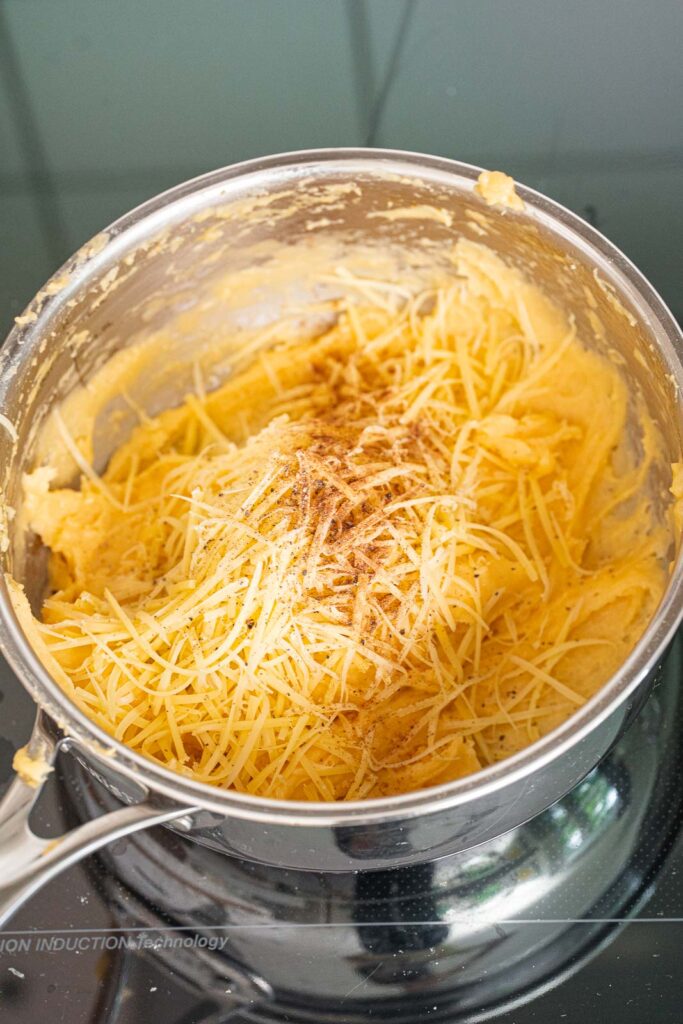 A stainless-steel saucepan containing mashed potatoes, topped with grated cheese and sprinkled with ground spices, sitting on a stovetop.