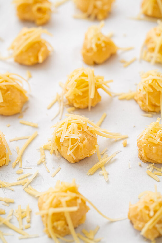 Small mounds of dough topped with grated cheese are arranged on a baking sheet lined with parchment paper.