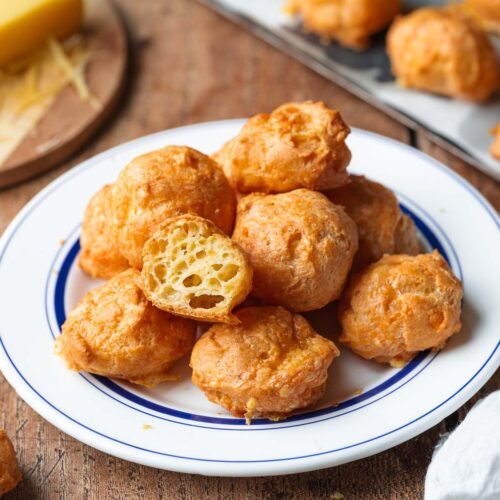 A plate of golden brown cheese puffs sits on a wooden table, with one puff broken open to show the airy interior. A block of cheese and more puffs are visible in the background.