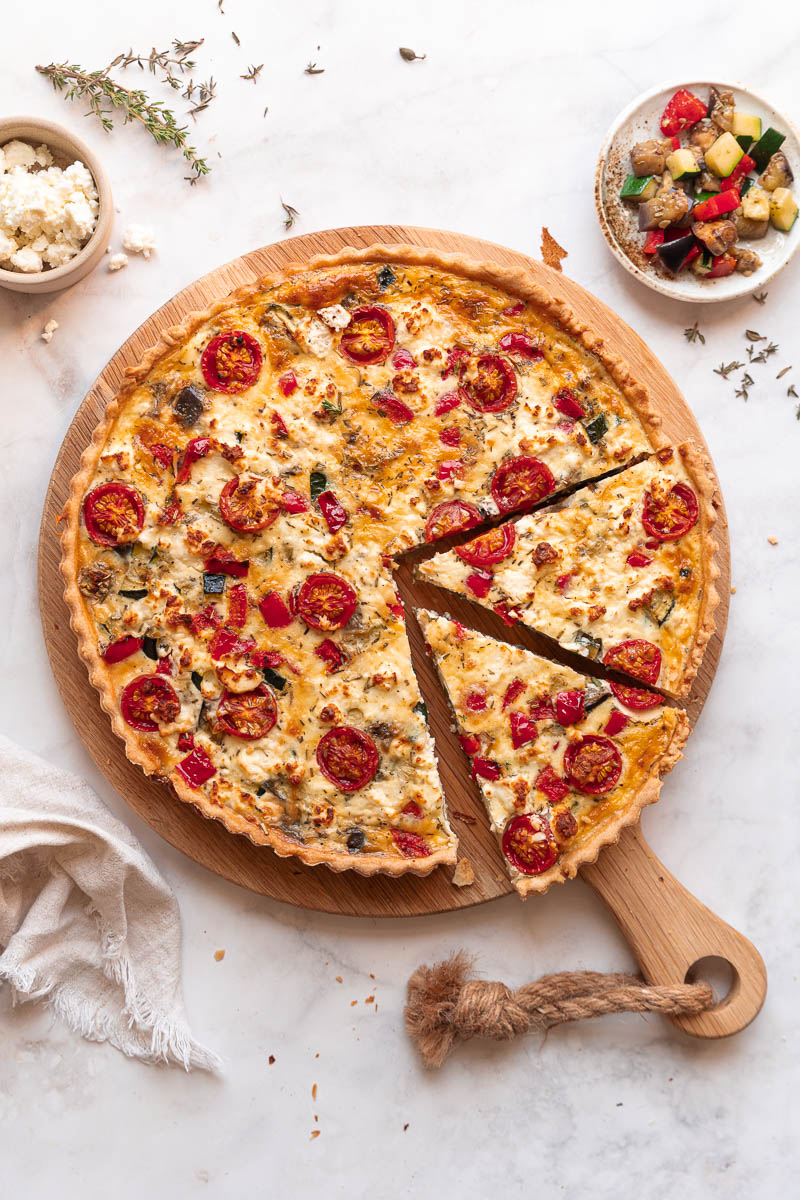 Eine Quiche mit Gemüse und Kirschtomaten ist auf einem Holzbrett in Scheiben geschnitten. Daneben stehen kleine Schälchen mit Feta und einer Gemüsemischung.