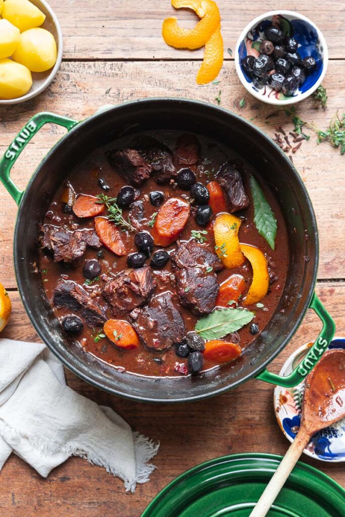 Une casserole de daube de bœuf à la provençale, agrémentée de carottes, d’olives noires, de feuilles de laurier et d’écorce d’orange, trône sur une table en bois, évoquant les saveurs de la cuisine provençale traditionnelle. À côté, un saladier de pommes de terre et une tasse d’olives noires viennent compléter ce tableau appétissant de daube provençale.
