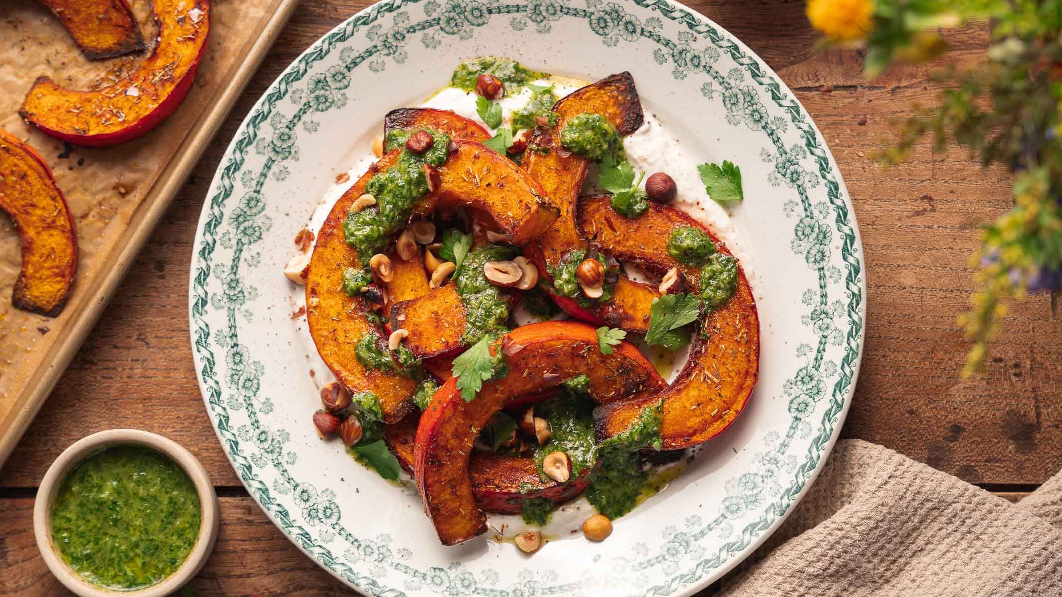 Des tranches de courge rôties disposées sur une assiette à motifs, nappées de sauce verte, d'herbes et de noix, avec de la sauce et d'autres morceaux de courge visibles à proximité sur une table en bois.