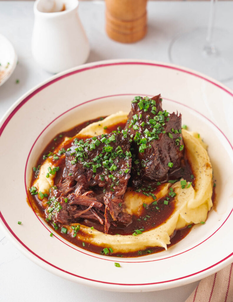 Côtes courtes braisées accompagnées de sauce et de purée de pommes de terre, garnies de ciboulette ciselée, servies dans un plat creux.
